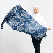 A model wearing a white hijab and black pants is holding a navy-blue batik scarf with a floral orchid design. The scarf is spread out in her hands, showcasing the design against a white background.