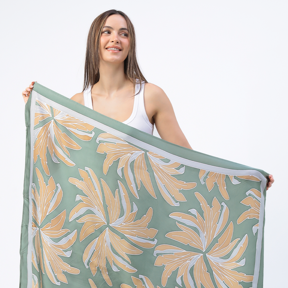 Woman showcasing the Moss Palma Scarf, a handcrafted batik scarf featuring an intricate palm-inspired design in soft green and beige tones. Made using traditional techniques, this versatile piece from Batik Boutique blends heritage craftsmanship with modern elegance.