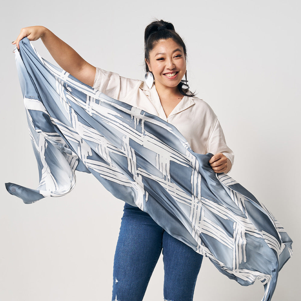 A woman is standing in front of white wall, showing grey brush color batik long scarf, handcrafted and artisan made locally in Malaysia