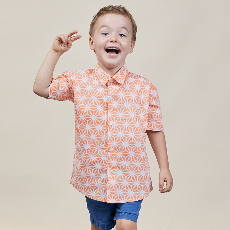A cheerful young boy posing with joy while wearing a Peach Firework patterned batik shirt, set against a neutral background