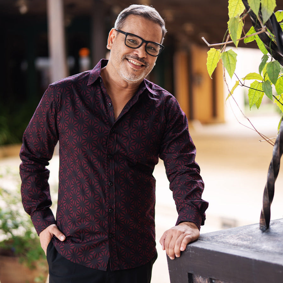 a man in a lifestyle photo leaning against a wooden structure  while wearing a batik shirt in the pattern garnet firework