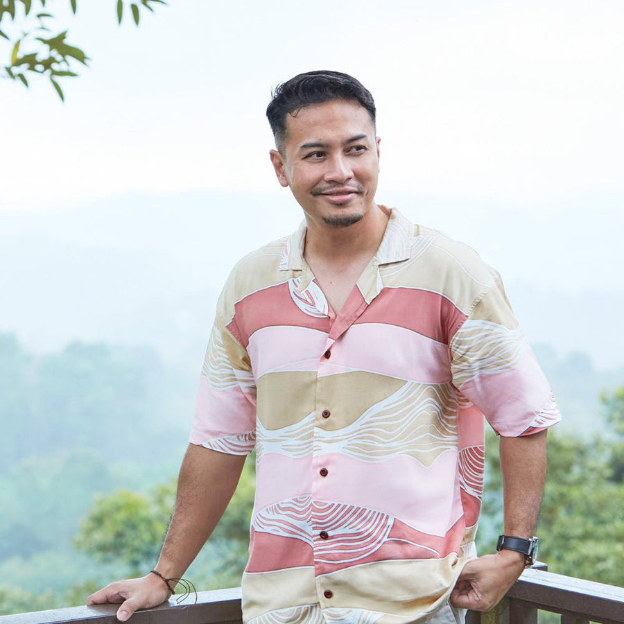 A man standing by the balcony with the background of forest. He's wearing batik shirt in pattern name Dawn Bukit
