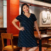 A women is standing beside a wood table and chair while posing in batik dress in jet tangga pattern. 