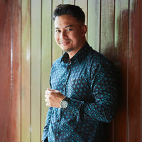 A man leaning standing in front of wood wall. He's wearing batik shirt in black alur. 