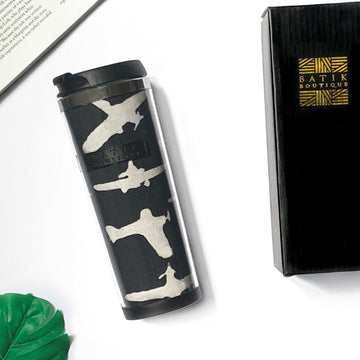 a photo of a black airplane tumbler made of batik against a neutral background with a tropical leaf, a black box from batik boutique and a book as decoration