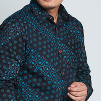 a close-up photo of man standing in front of a white wall wearing a long-sleeved batik shirt in black alur