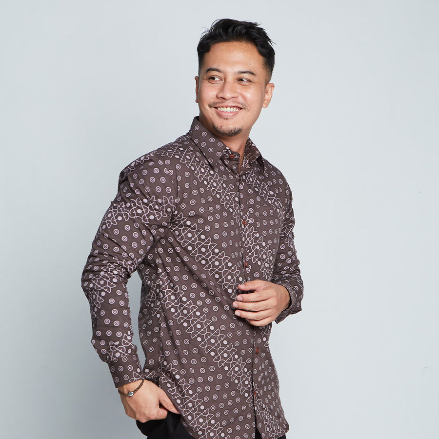 a man posing in front of a white wall while wearing a long-sleeved batik shirt in brown alur