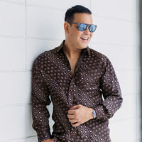 a man posing in front of a white brick wall with shades and wearing a batik shirt in brown alur
