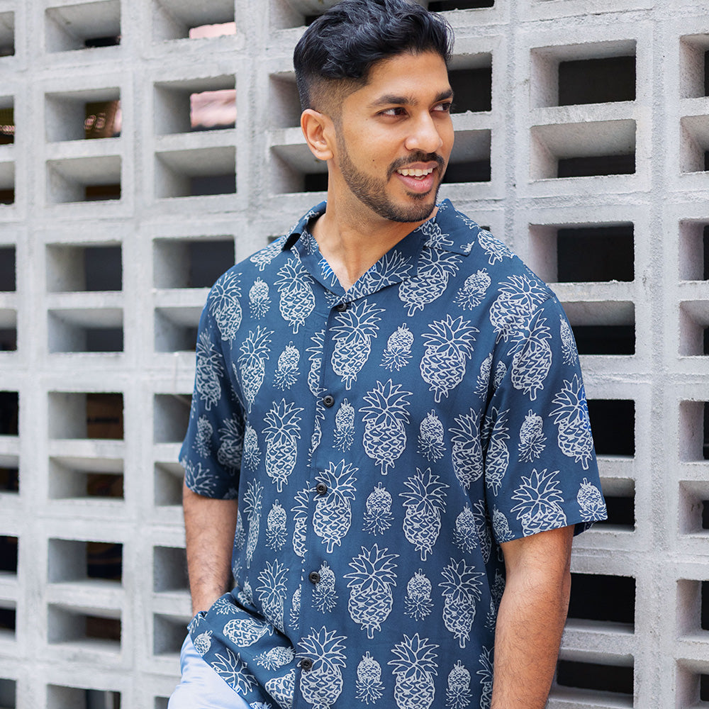 a lifestyle photo of man leaning by wall of brick wearing batik cuban shirt in navy pineapple pattern