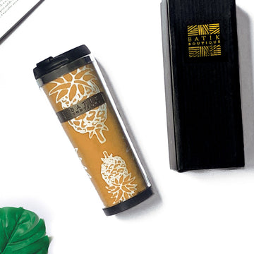 a lifestyle photo of a golden pineapple tumbler made of batik against a neutral background with a black box from batik boutique, a tropical leaf and a book as decorations