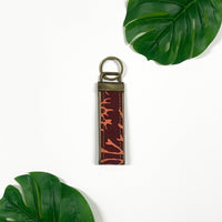a lifestyle photo of a keyfob made of batik in the pattern maroon coral against a neutral background surrounded by two tropical plants