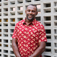A man is wearing batik short sleeve shirt in Crimson Nasi Lemak, a red color cotton batik shirt in Nasi Lemak motif. Standing and leaning on brick wall