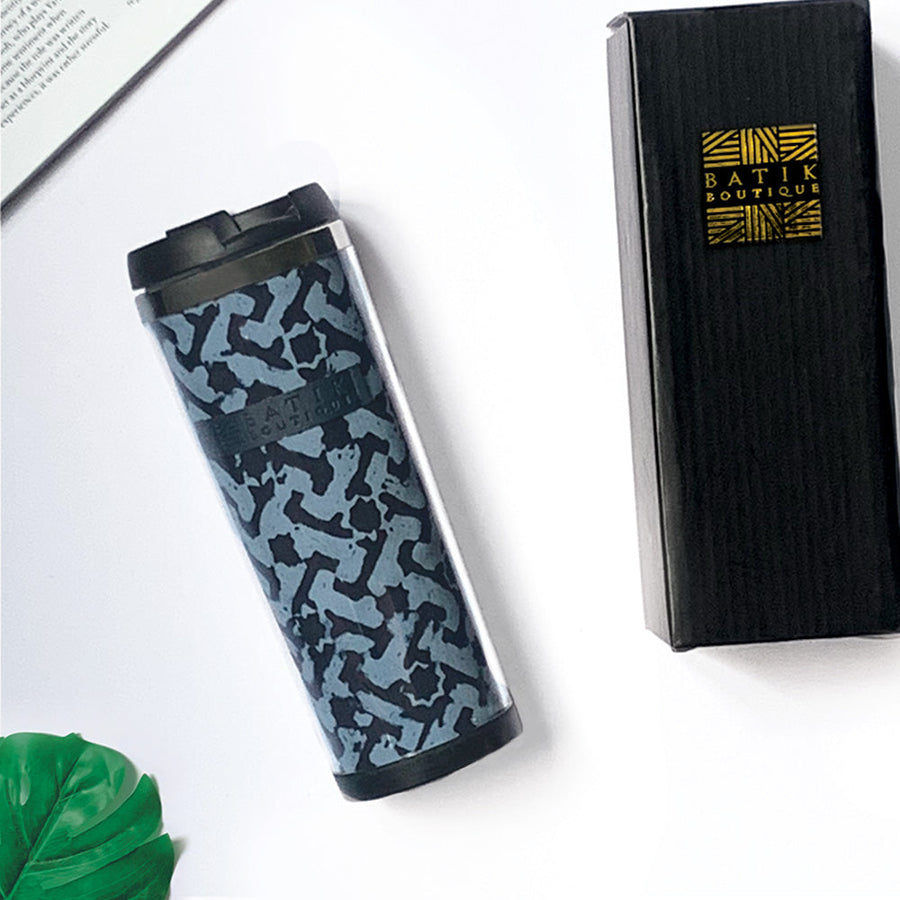 a lifestyle photo of a batik tumbler in the pattern midnight arabesque made of batik against a neutral background with a tropical leaf, black box and a book as decorations
