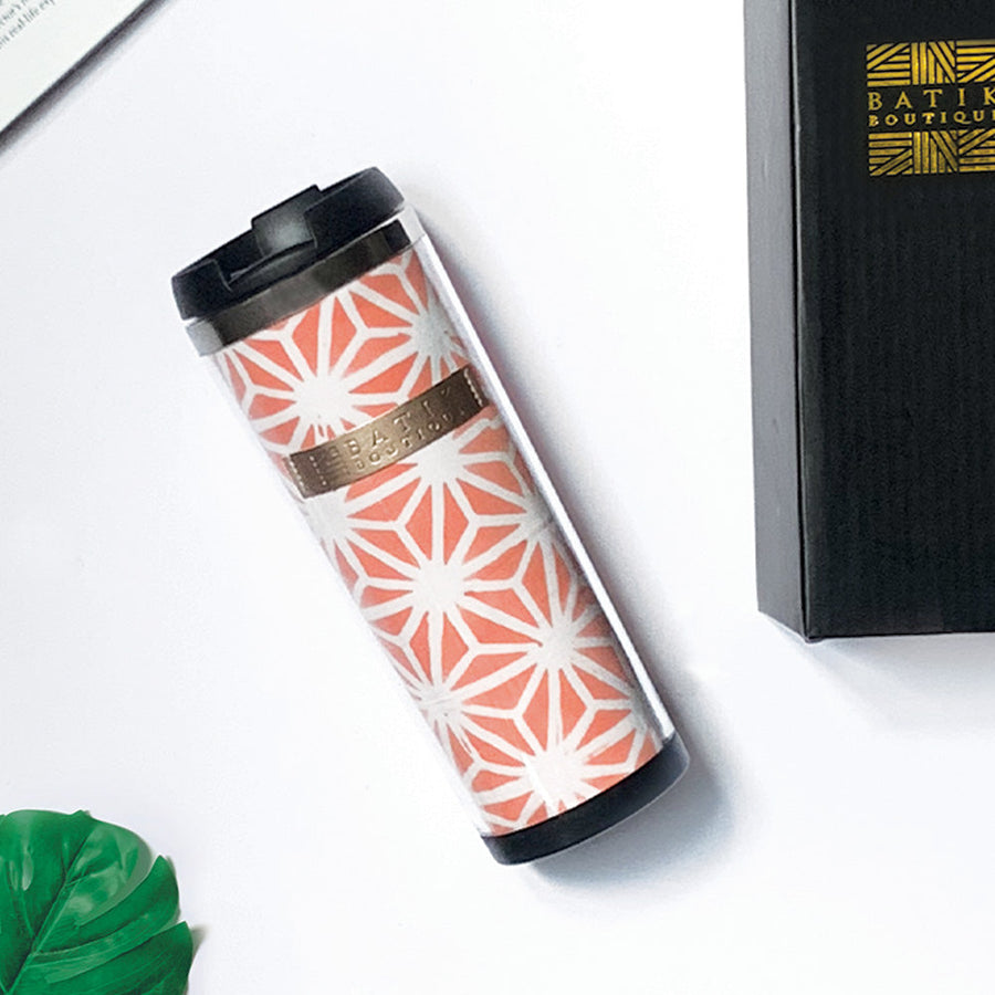 a batik tumbler in the pattern peach firework laying on a white surface surrounded by a black box with the batik boutique logo, a leaf and a book
